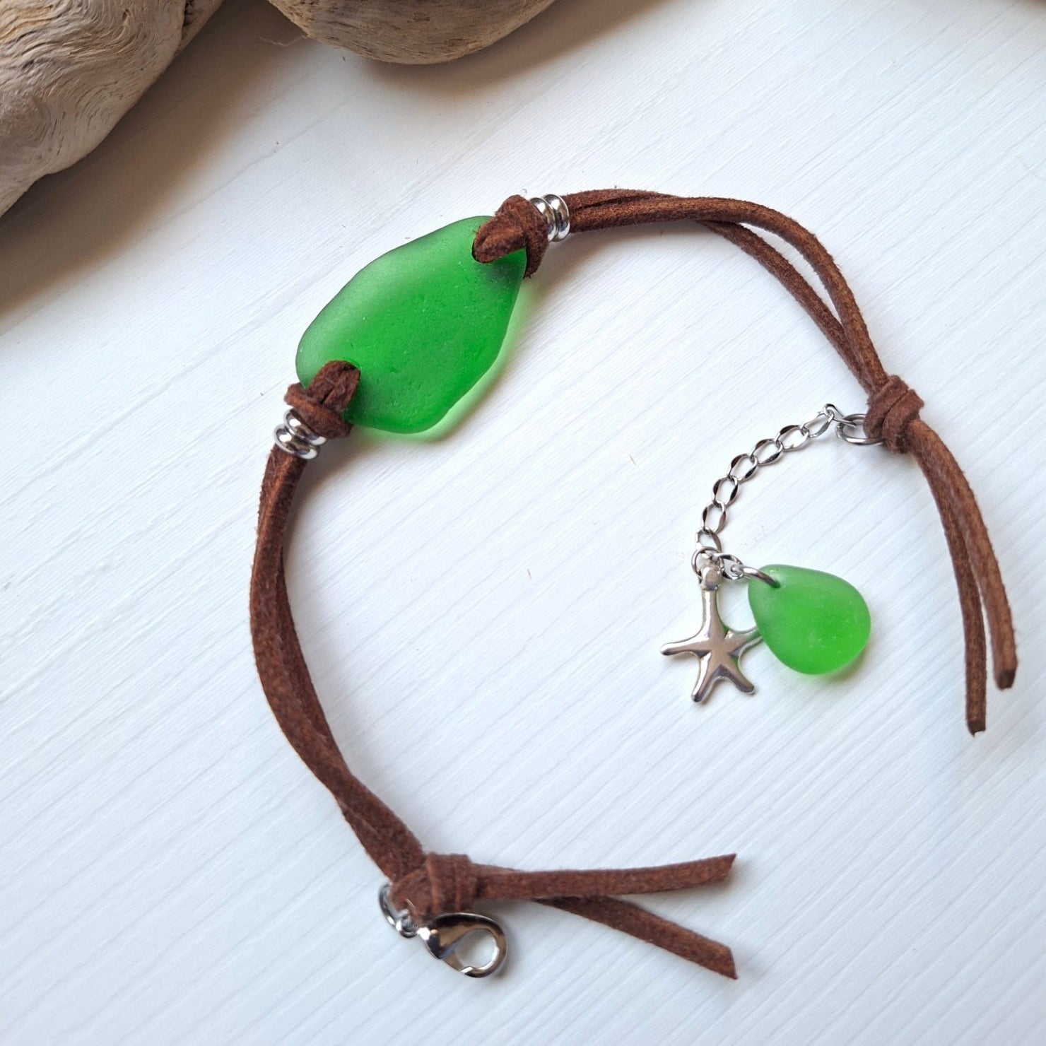 A bohemian-style sea glass bracelet with a brown faux suede cord, featuring a large green pieece of sea glass, a smaller green sea glass charm, a silver starfish charm, and stainless steel spacer beads.
