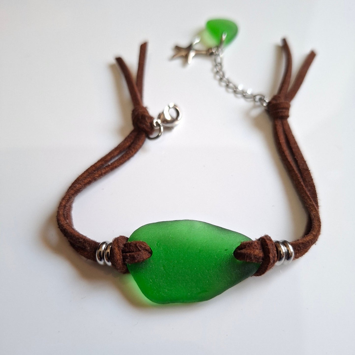 A bohemian-style sea glass bracelet with a brown faux suede cord, featuring a large green pieece of sea glass, a smaller green sea glass charm, a silver starfish charm, and stainless steel spacer beads.