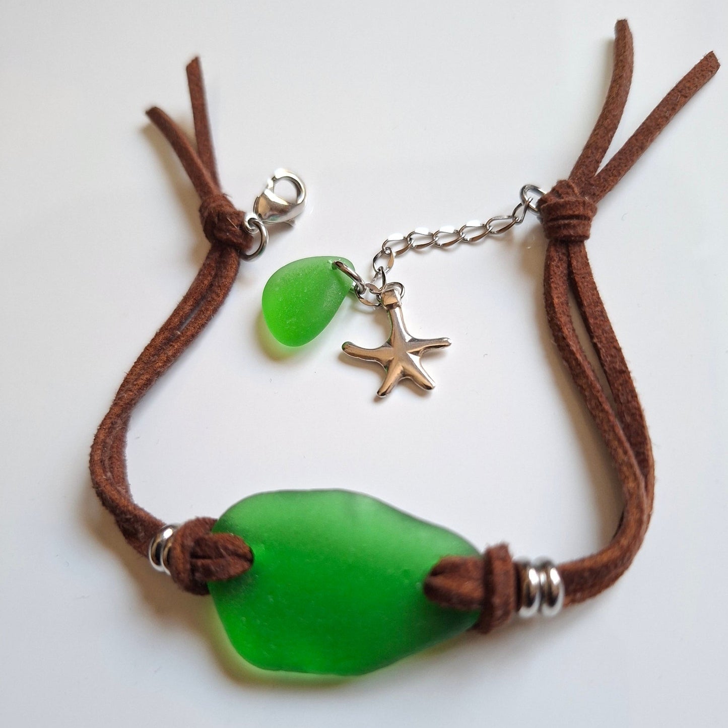 A bohemian-style sea glass bracelet with a brown faux suede cord, featuring a large green pieece of sea glass, a smaller green sea glass charm, a silver starfish charm, and stainless steel spacer beads.