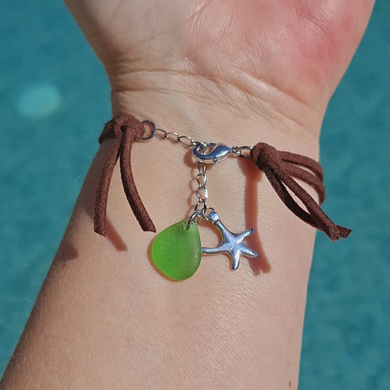 A bohemian-style sea glass bracelet with a brown faux suede cord, featuring a large green pieece of sea glass, a smaller green sea glass charm, a silver starfish charm, and stainless steel spacer beads.