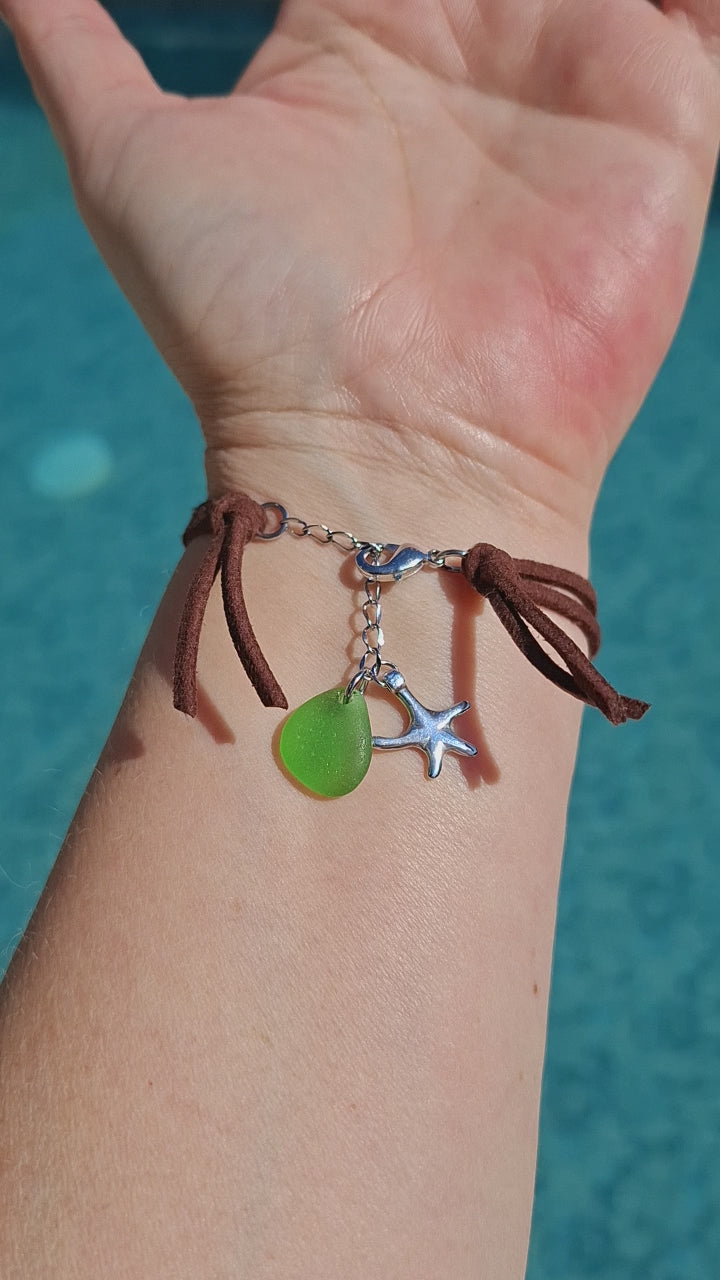 A bohemian-style sea glass bracelet with a brown faux suede cord, featuring a large green pieece of sea glass, a smaller green sea glass charm, a silver starfish charm, and stainless steel spacer beads.